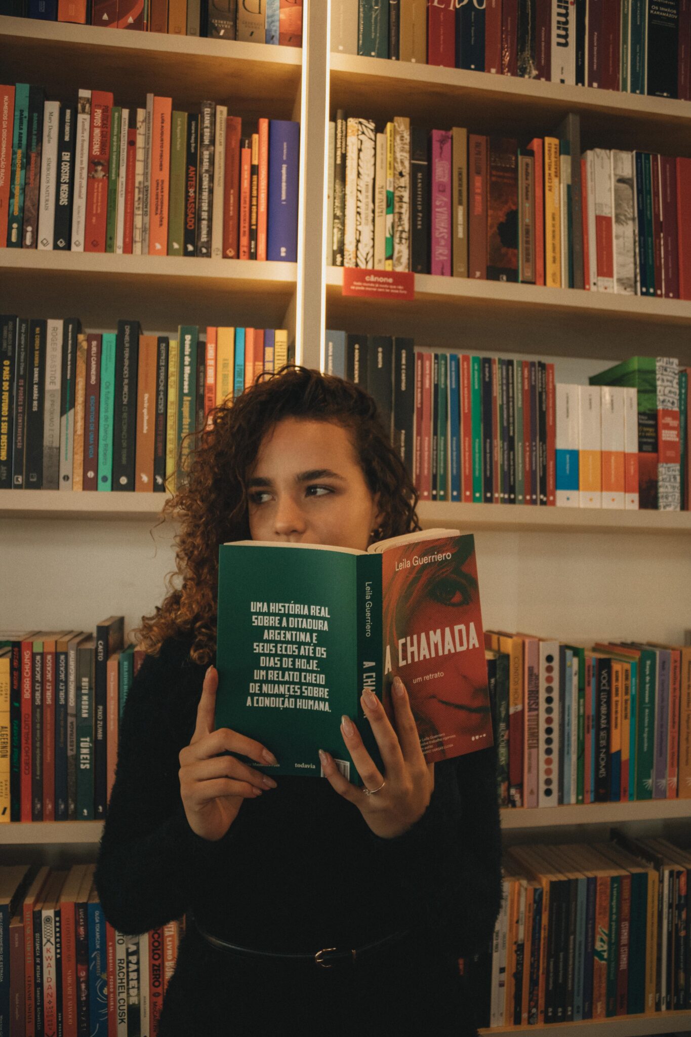 Woman reading a paperback book in a cozy library setting, surrounded by bookshelves.