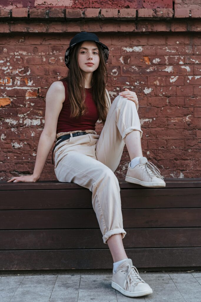 Stylish teenager in a bucket hat and tank top sitting relaxed against an urban brick wall.